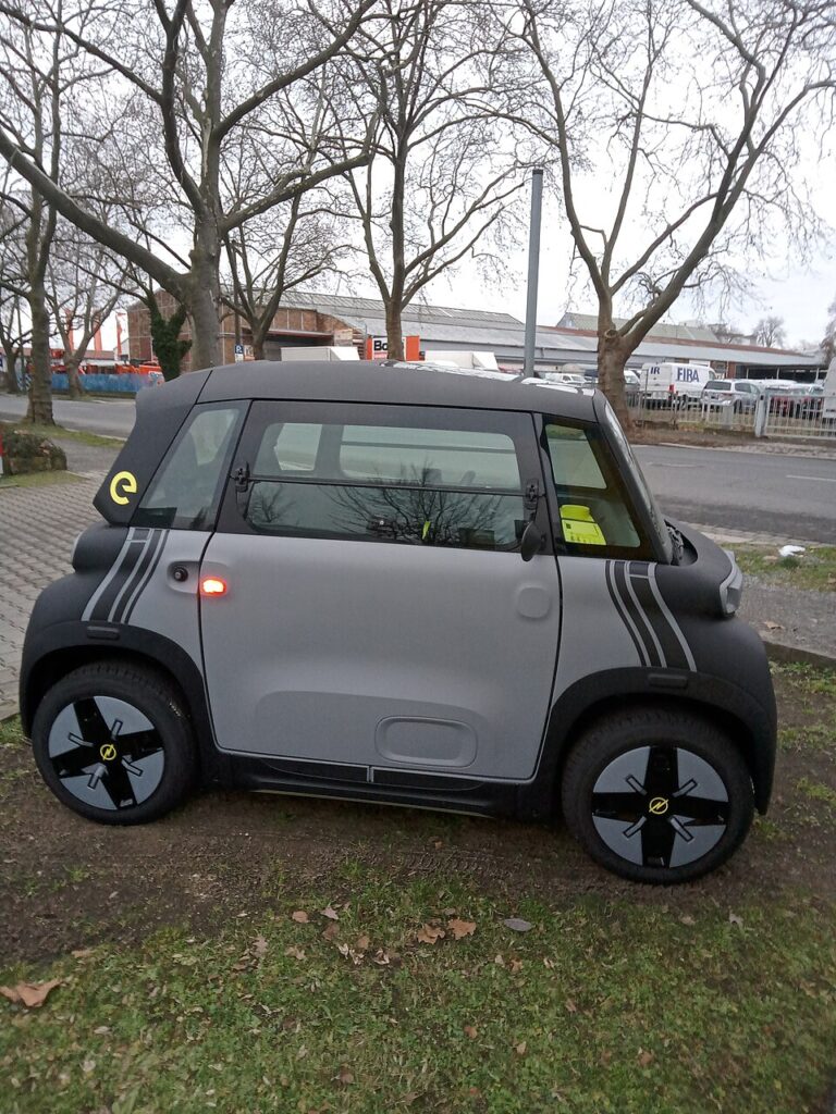 Microcar electric cu caroserie închisă, gri cu negru, parcat pe iarbă, văzut din lateral