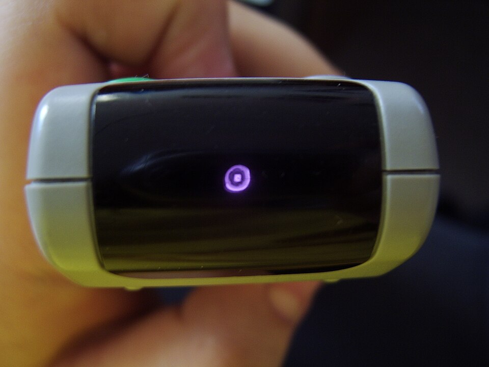 Capătul unei telecomenzi ținute în mână, cu ledul infraroșu care apare ca un punct violet luminos pe ecranul camerei